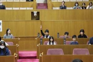 こども議会体験会でこども（2人目）が議員席で手を挙げている写真