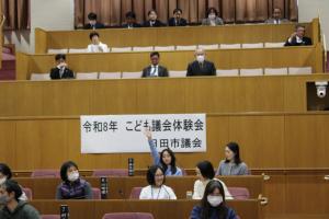こども議会体験会でこども（5人目）が議員席で手を挙げている写真