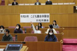 こども議会体験会でこども（6人目）が議員席で手を挙げている写真