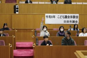 こども議会体験会でこども（7人目）が議員席で手を挙げている写真