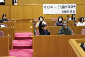 こども議会体験会でこども（8人目）が議員席で手を挙げている写真