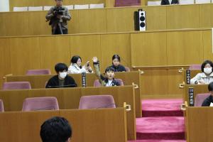 こども議会体験会でこども（10人目）が議員席で手を挙げている写真